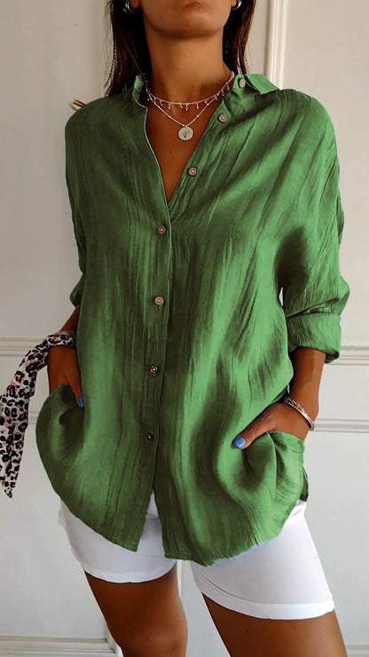 Frau trägt grüne Leinenbluse mit Knopfleiste und weißen Hosen. Modischer Look, ideal für Sommer. Accessoires: Halskette, Armband, gemusterter Schal.