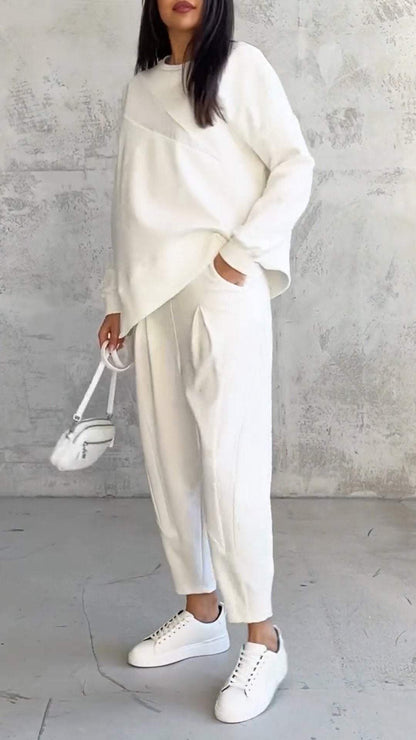 Frau in weißem Oversize-Pullover und Jogginghose, weiße Sneakers, kleine weiße Handtasche, vor grauer Betonwand. Minimalistischer Modestil.