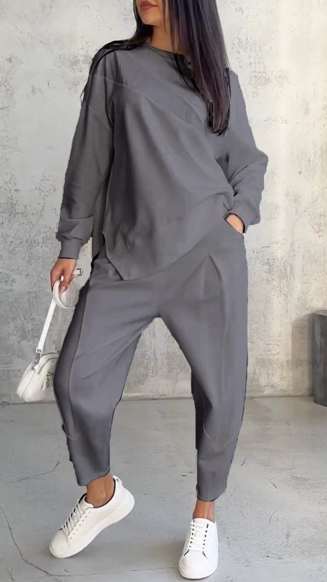 Frau in grauem Oversize-Sweatshirt und Jogginghose, weiße Sneakers, weiße Handtasche, lässiger Look, modische Freizeitkleidung, Betonhintergrund.