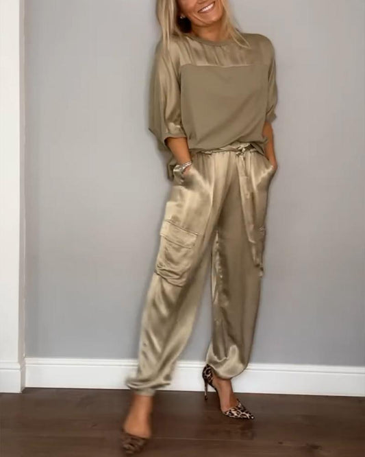Frau in eleganter, beiger Satin-Hose und passendem Oberteil, posiert lächelnd vor grauer Wand. Modetrend, Damenmode, Satin-Outfit.