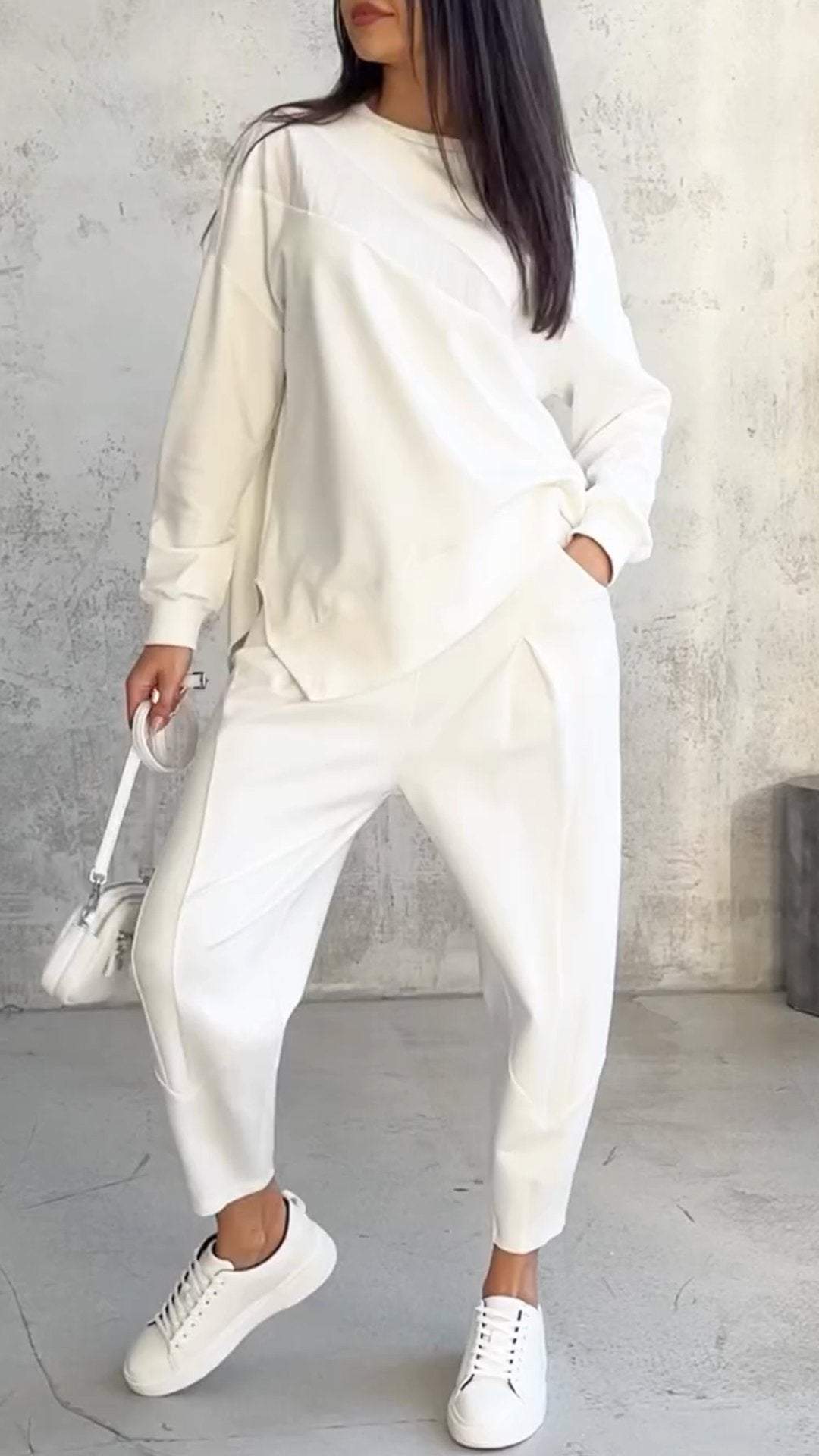 Frau in weißem Oversize-Sweatshirt und Jogginghose, weiße Sneakers, hält weiße Handtasche. Modetrend, lässiger Look, Streetstyle.