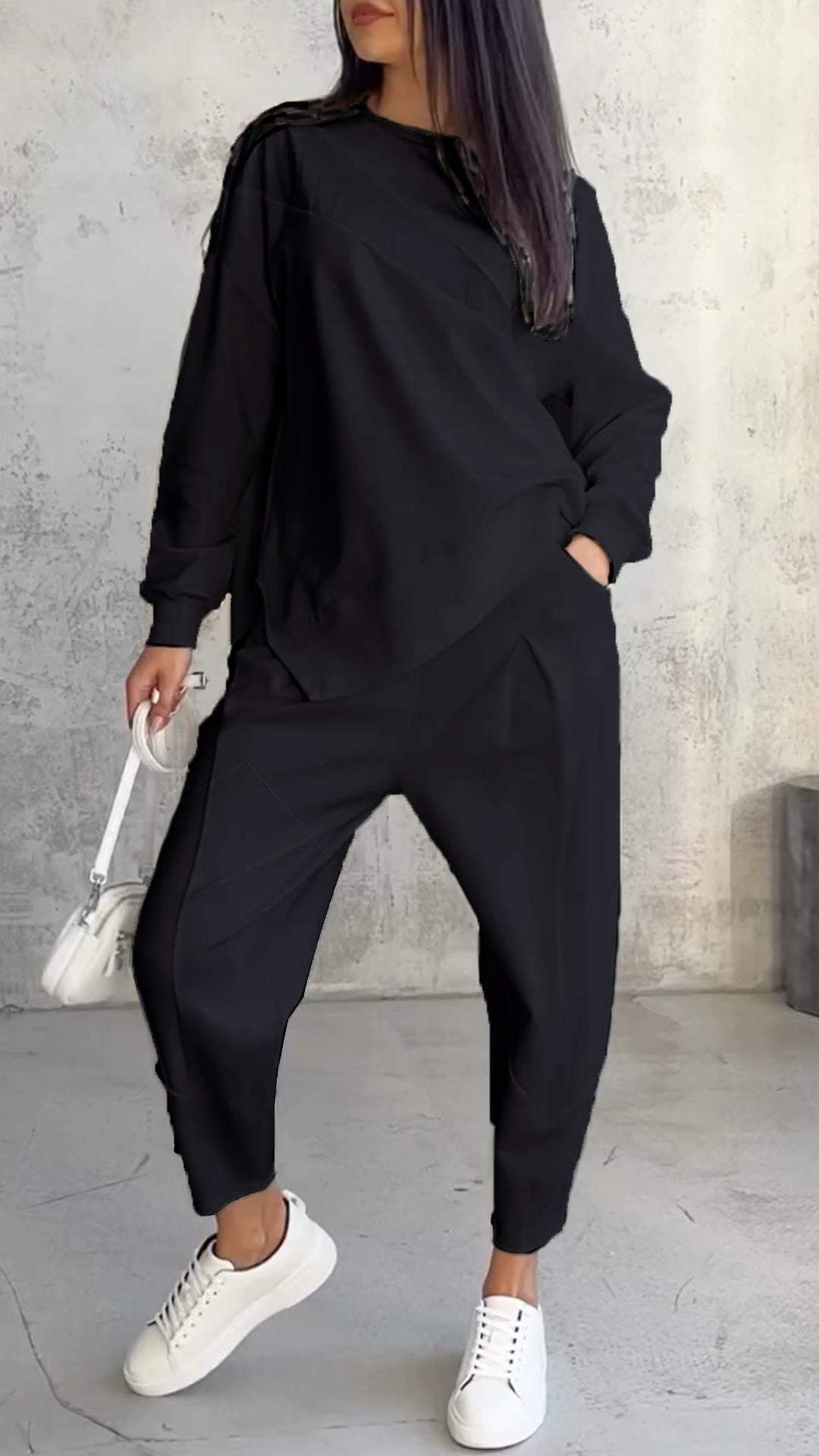 Frau in schwarzem Oversize-Sweatshirt und Jogginghose, weiße Sneakers, weiße Handtasche, grauer Betonhintergrund. Modetrend, lässiger Stil.