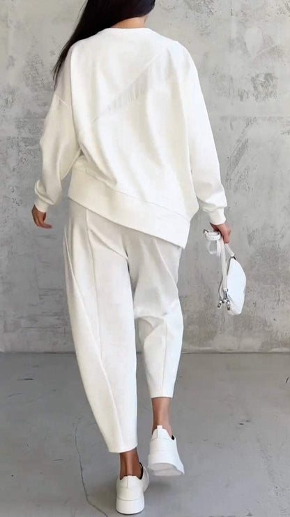 Frau in weißem Oversize-Sweatshirt und Jogginghose, trägt weiße Sneakers und kleine Handtasche, vor grauer Betonwand. Fashion, Streetstyle, Loungewear.