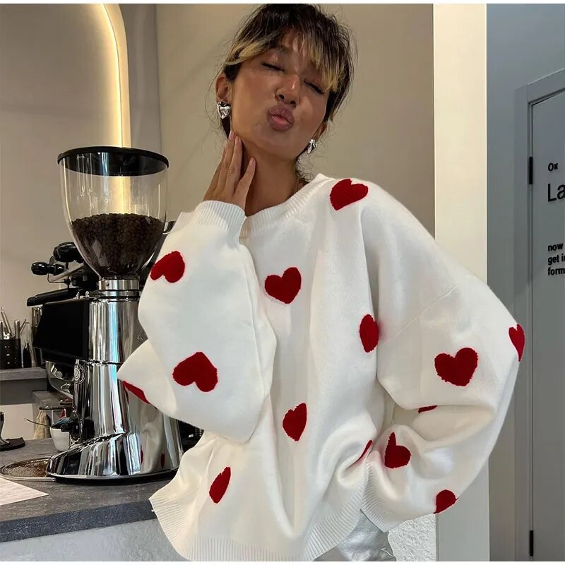 Frau in weißem Pullover mit roten Herzen posiert vor einer Kaffeemaschine. Modetrend, lässiger Stil, gemütlicher Look, Café-Hintergrund.