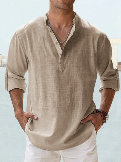 Mann trägt beiges Leinenhemd mit Stehkragen und Knopfleiste, kombiniert mit weißen Hosen. Lässige Sommermode, ideal für Strand oder Freizeit.
