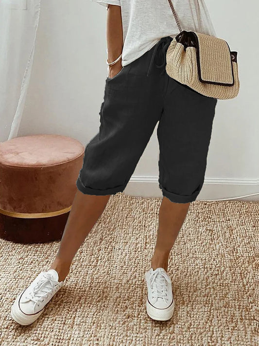 Frau in lässiger Sommermode mit weißem T-Shirt, schwarzen Bermuda-Shorts und weißen Sneakers. Strohtasche als Accessoire. Perfekt für Freizeit und Alltag.