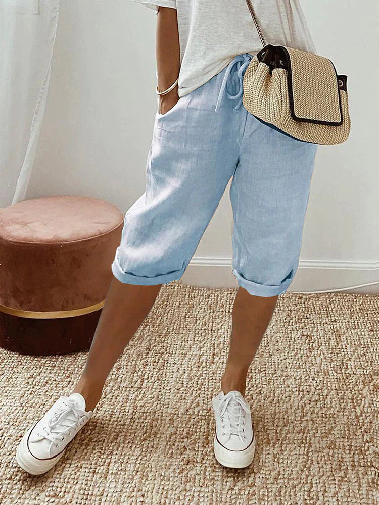 Frau in lässiger Sommermode: Hellblaue Leinen-Shorts, weißes T-Shirt, weiße Sneaker, Strohtasche. Perfekt für Freizeit und Strand.