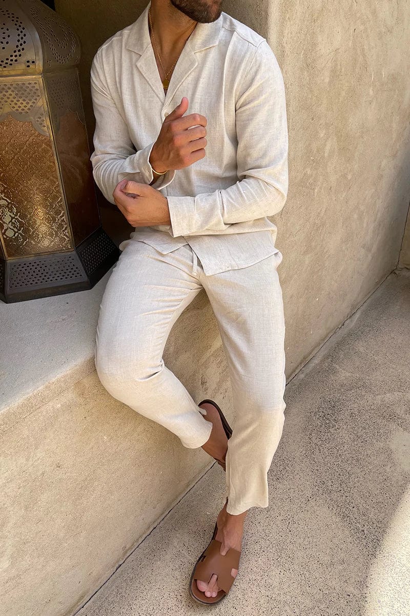 Mann in beigem Leinenanzug sitzt entspannt, trägt braune Sandalen. Stilvolle Sommermode, lässiger Look, ideal für warme Tage.