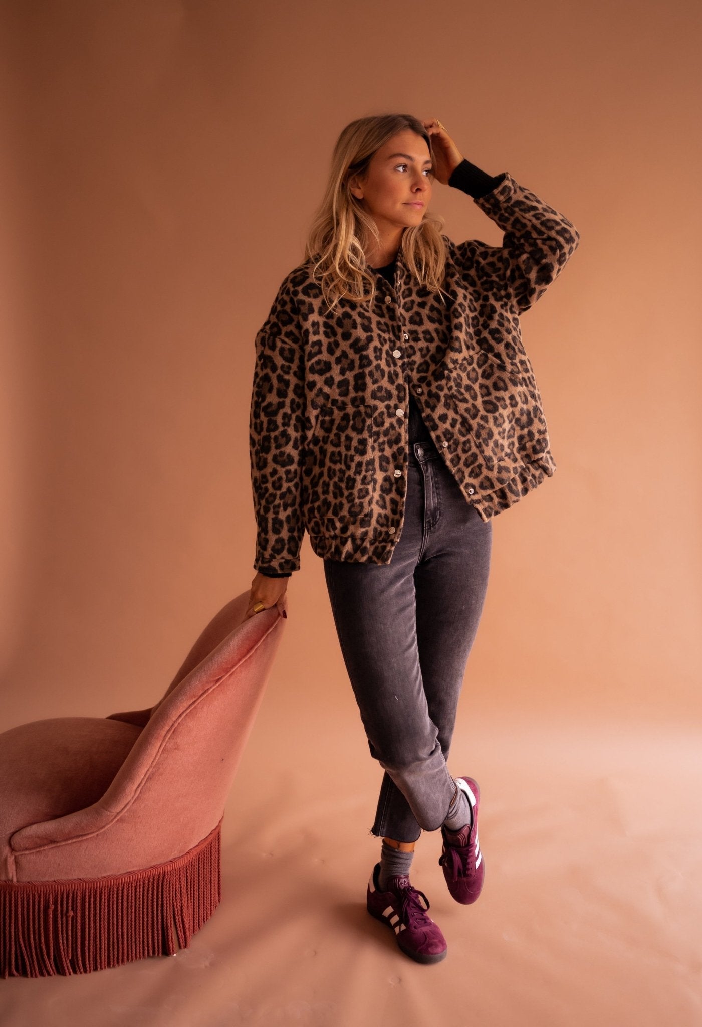 Frau in Leopardenjacke, grauer Jeans und lila Sneakers lehnt an rosa Sessel. Mode, Herbstlook, trendige Damenbekleidung, stilvolles Outfit.