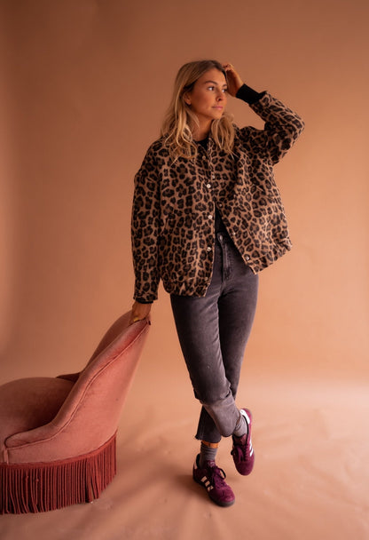 Frau in Leopardenjacke, grauer Jeans und lila Sneakers lehnt an rosa Sessel. Mode, Herbstlook, trendige Damenbekleidung, stilvolles Outfit.