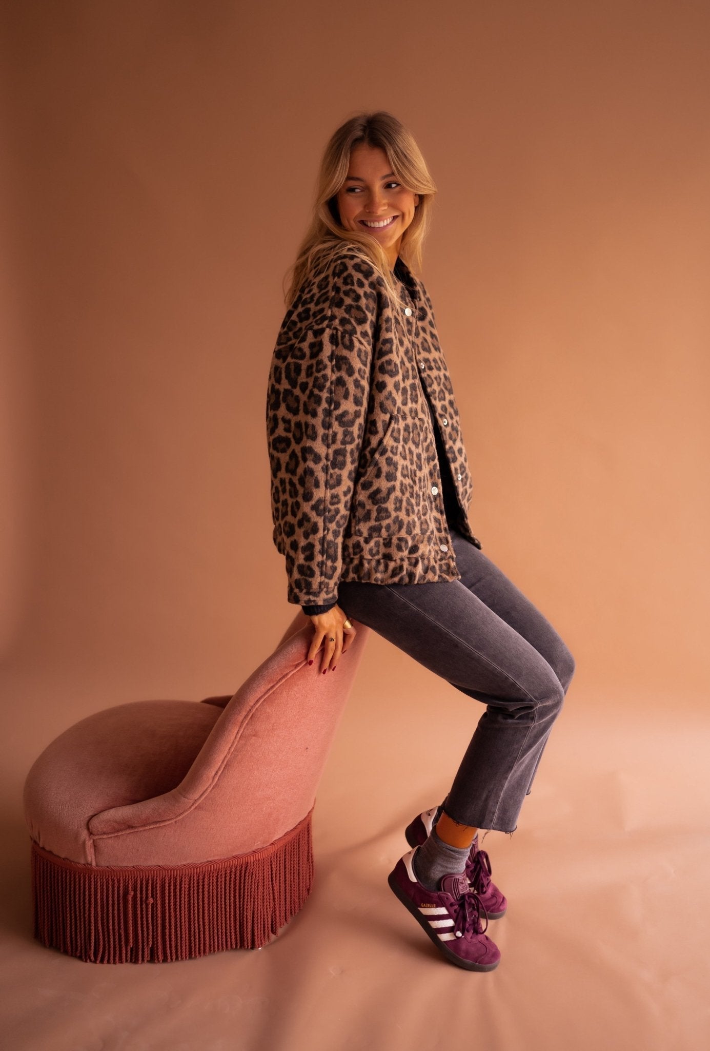 Frau in Leopardenjacke sitzt auf rosa Sessel, trägt graue Jeans und lila Adidas-Sneaker. Mode, Herbstlook, trendige Damenbekleidung.