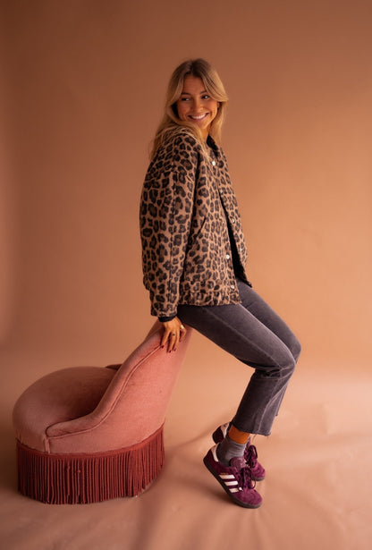 Frau in Leopardenjacke sitzt auf rosa Sessel, trägt graue Jeans und lila Adidas-Sneaker. Mode, Herbstlook, trendige Damenbekleidung.