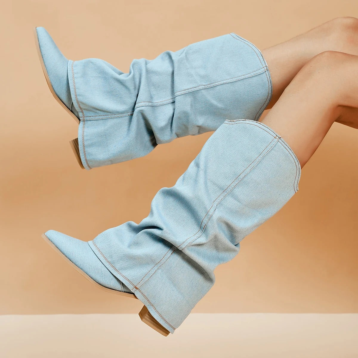 Hellblaue Denim-Stiefel mit weitem Schaft und spitzer Zehenpartie, getragen von Modellbeinen vor beigem Hintergrund. Modetrend, Damenstiefel, Jeanslook.