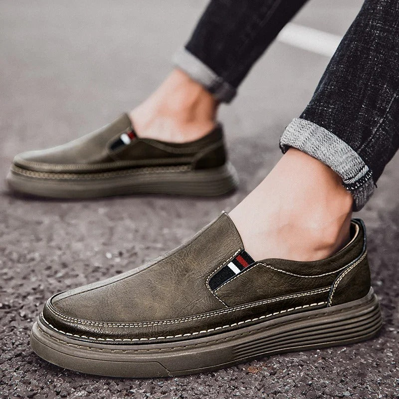 Graue Herren-Slipper aus Leder mit gestreiftem Detail, bequeme Freizeitschuhe auf Asphalt. Modische, rutschfeste Schuhe für den Alltag.