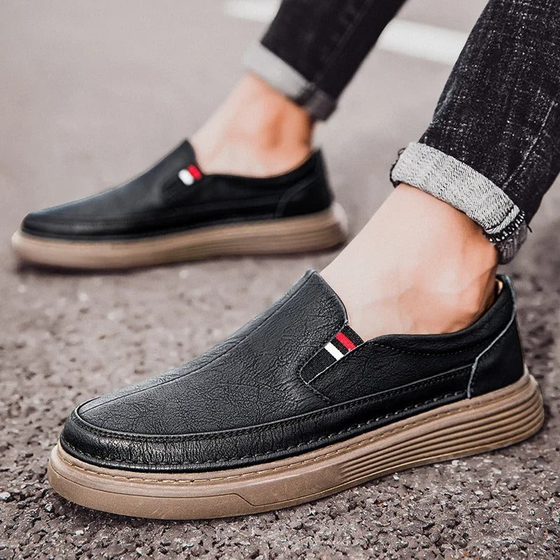 Schwarze Herren-Slipper aus Leder mit brauner Sohle, getragen auf Asphalt. Modische Freizeitschuhe, ideal für den Alltag.