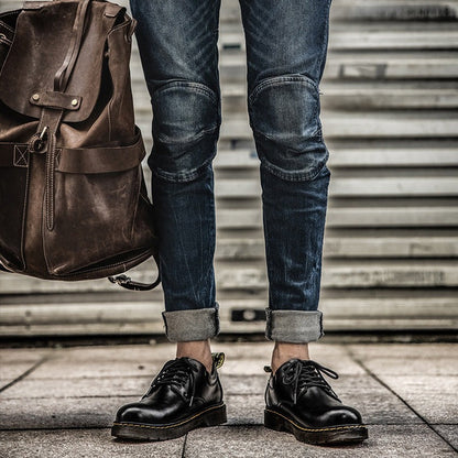 Mann mit hochgekrempelten Jeans, schwarzen Lederschuhen und braunem Lederrucksack vor Metalltor. Urbaner Stil, modische Herrenaccessoires.