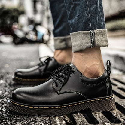 Schwarze Lederhalbschuhe mit gelber Naht, getragen von Person in hochgekrempelten Jeans, auf städtischem Gehweg. Modische Herrenschuhe, urbaner Stil.