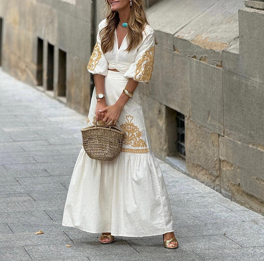 Frau in weißem Maxikleid mit goldenen Stickereien, hält einen geflochtenen Korb. Sommermode, eleganter Boho-Stil, Stadtstraße.