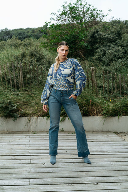 Frau in blauer Jeans und gemustertem Pullover steht auf Holzsteg vor grüner Naturkulisse. Mode, Freizeitkleidung, Outdoor-Fotografie.