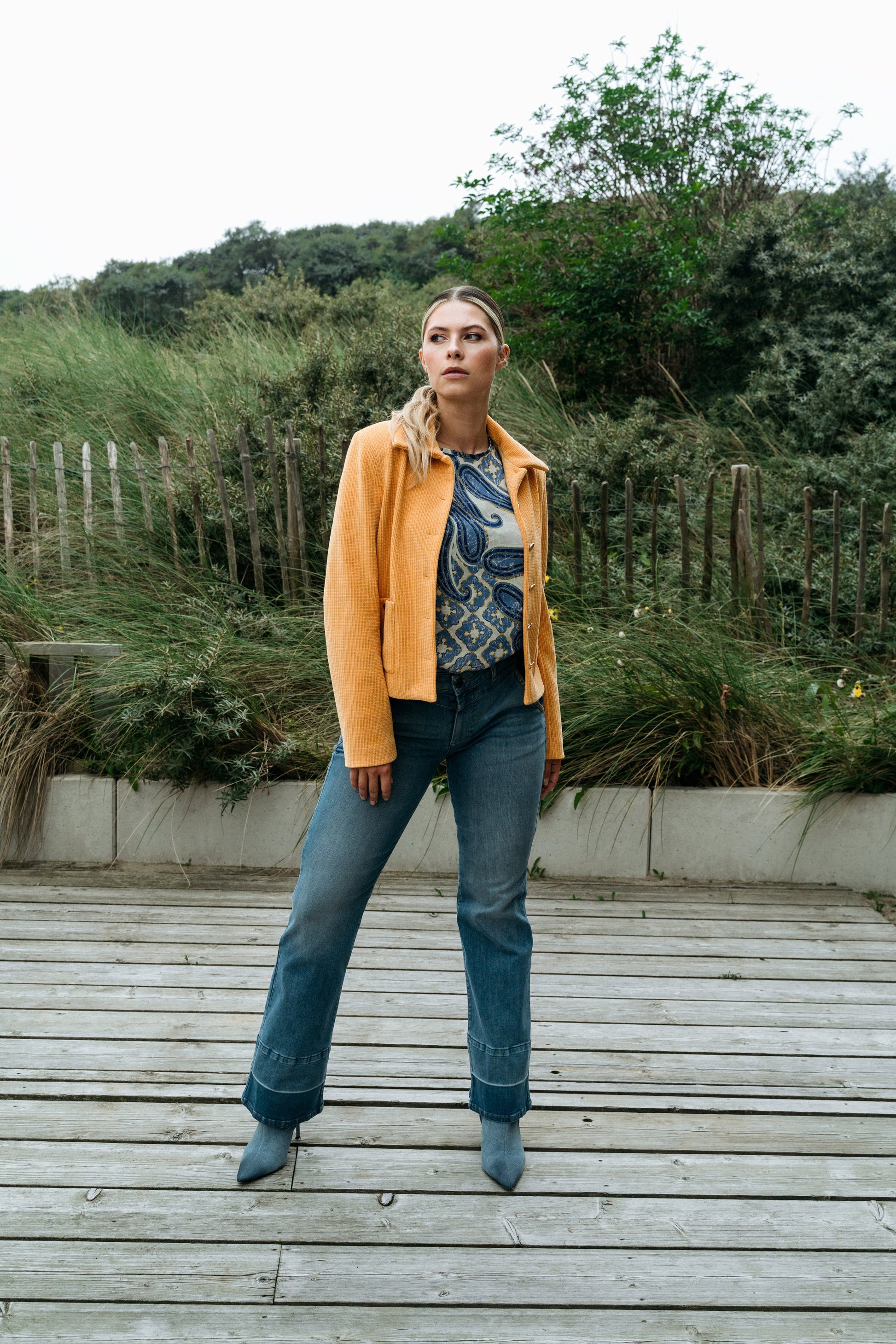 Frau in gelber Jacke und blauer Jeans steht auf Holzsteg vor grüner Landschaft. Modetrend, Freizeitkleidung, Outdoor-Fashion.