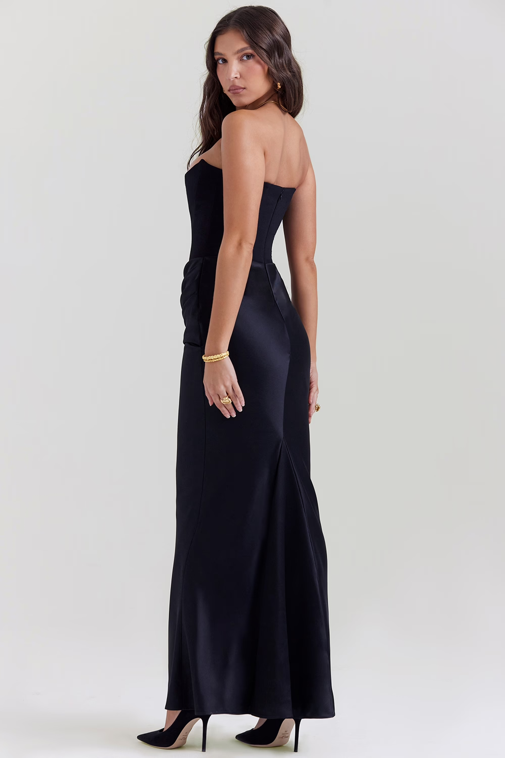 Frau in elegantem, schwarzem Abendkleid, schulterfrei, mit goldenen Armbändern und schwarzen High Heels. Perfekt für formelle Anlässe und Abendveranstaltungen.