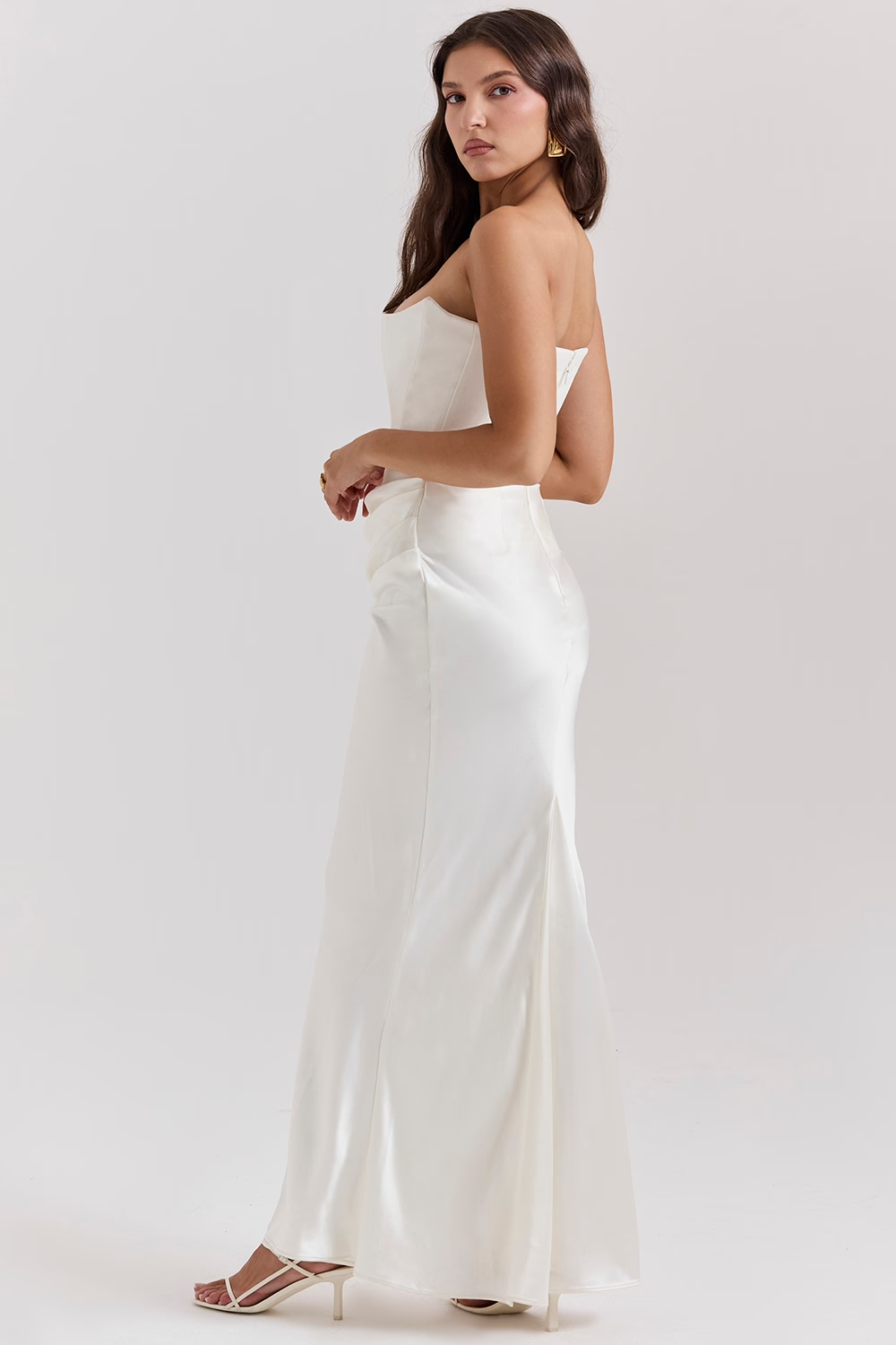 Frau in elegantem, schulterfreiem weißen Abendkleid, seitlich stehend, mit langen braunen Haaren und weißen High Heels, vor neutralem Hintergrund.