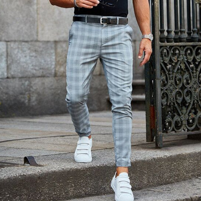 Mann in grauer karierter Hose und weißen Sneakers, geht auf einer Steinpflasterstraße. Elegante Herrenmode, lässiger Stil, urbane Umgebung.