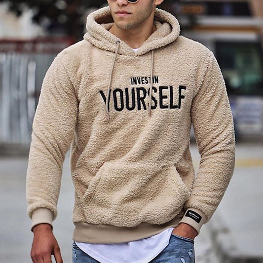 Mann in beigem Fleece-Hoodie mit Aufschrift "Invest in Yourself", lässiger Stil, Jeans, urbaner Hintergrund. Ideal für Mode- und Streetwear-Themen.
