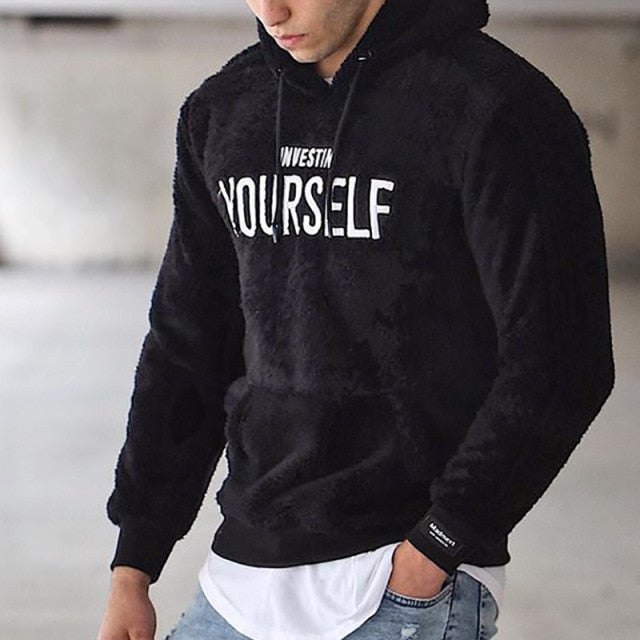 Mann trägt schwarzen Hoodie mit "Invest in Yourself"-Aufdruck, lässiger Stil, Jeans, urbaner Hintergrund. Modetrend, Herrenbekleidung, Streetwear.
