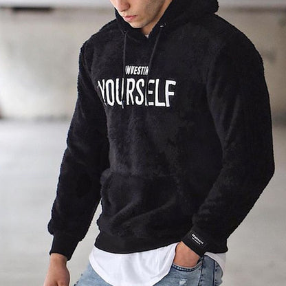 Mann trägt schwarzen Hoodie mit "Invest in Yourself"-Aufdruck, lässiger Stil, Jeans, urbaner Hintergrund. Modetrend, Herrenbekleidung, Streetwear.