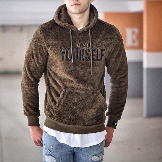Junger Mann in braunem Fleece-Hoodie mit "Invest in Yourself"-Aufdruck, lässig kombiniert mit zerrissenen Jeans in urbaner Umgebung.