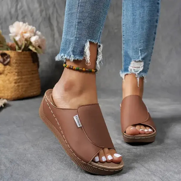 Damenfüße in braunen Sandalen mit Keilabsatz und offener Spitze, getragen zu zerrissenen Jeans. Hintergrund: Korb mit Blumen.