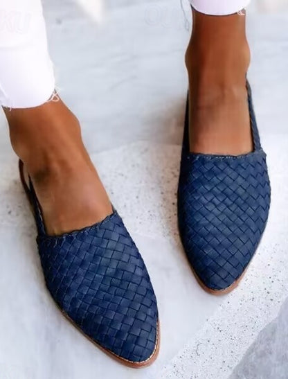 Damenfüße in blauen, geflochtenen Slippern auf grauem Boden. Modische Sommerschuhe, lässiger Stil, ideal für warme Tage.