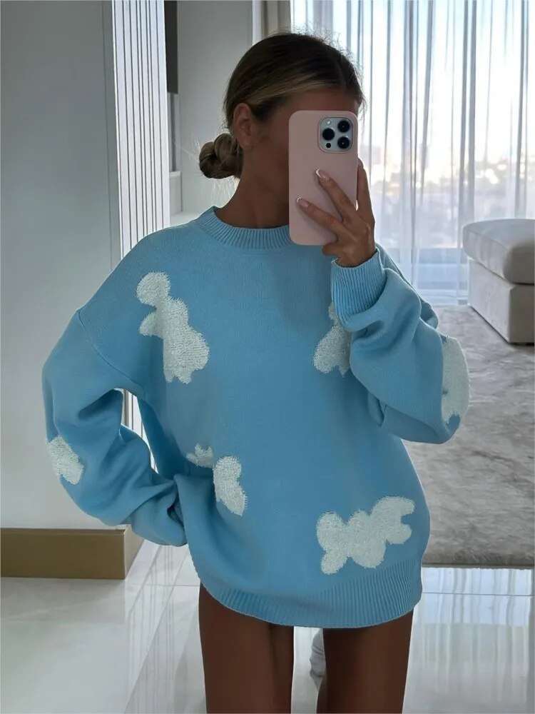Frau in blauem Pullover mit weißen Wolkenmuster, hält Smartphone vor Gesicht. Modernes Wohnzimmer im Hintergrund. Trendige Mode, gemütlicher Stil.