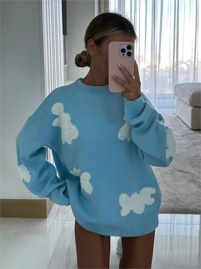 Frau in blauem Pullover mit weißen Wolkenmuster, hält Smartphone vor Gesicht. Modernes Wohnzimmer im Hintergrund. Trendige Mode, gemütlicher Stil.