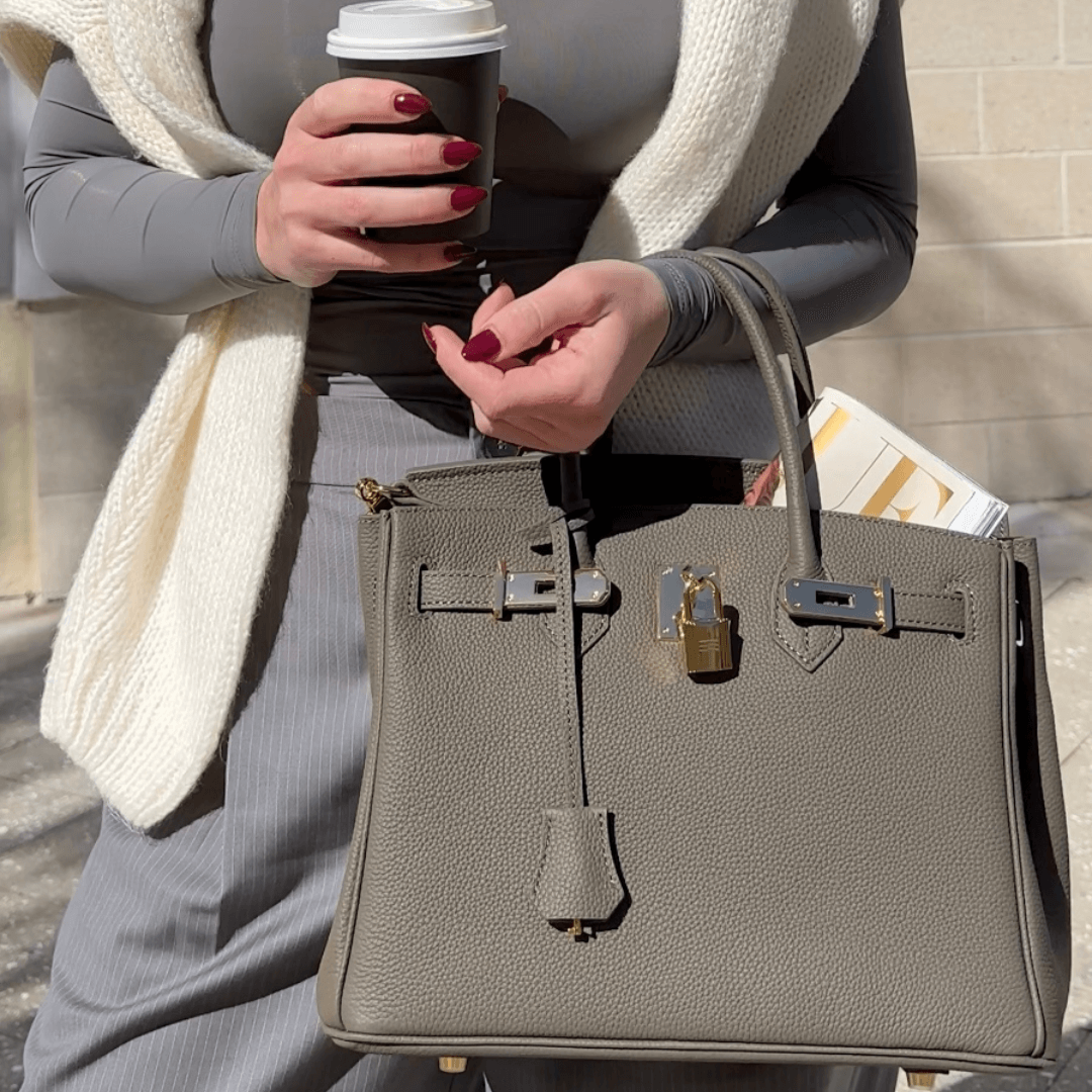 Frau mit grauer Birkin-Tasche, Kaffeebecher und Zeitschrift. Elegante Mode, Luxusaccessoire, Streetstyle.