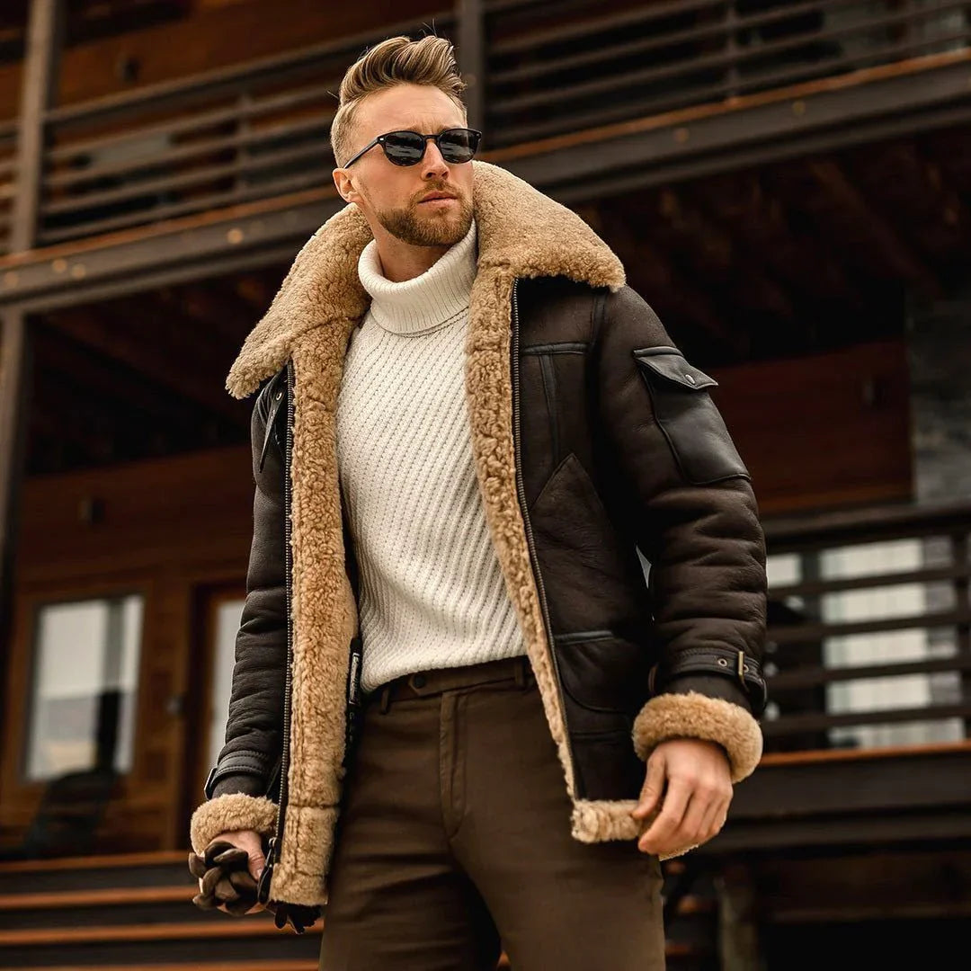 Mann in brauner Lederjacke mit Fellkragen, weißem Rollkragenpullover und Sonnenbrille vor Holzhaus. Wintermode, Herrenbekleidung, stilvoll.