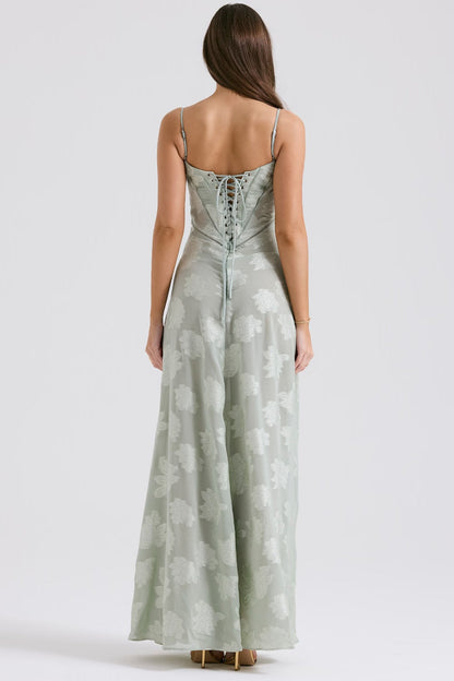 Frau in langem, mintgrünem Abendkleid mit Blumenmuster und Schnürung am Rücken, stehend vor neutralem Hintergrund. Elegantes Design, Abendmode.