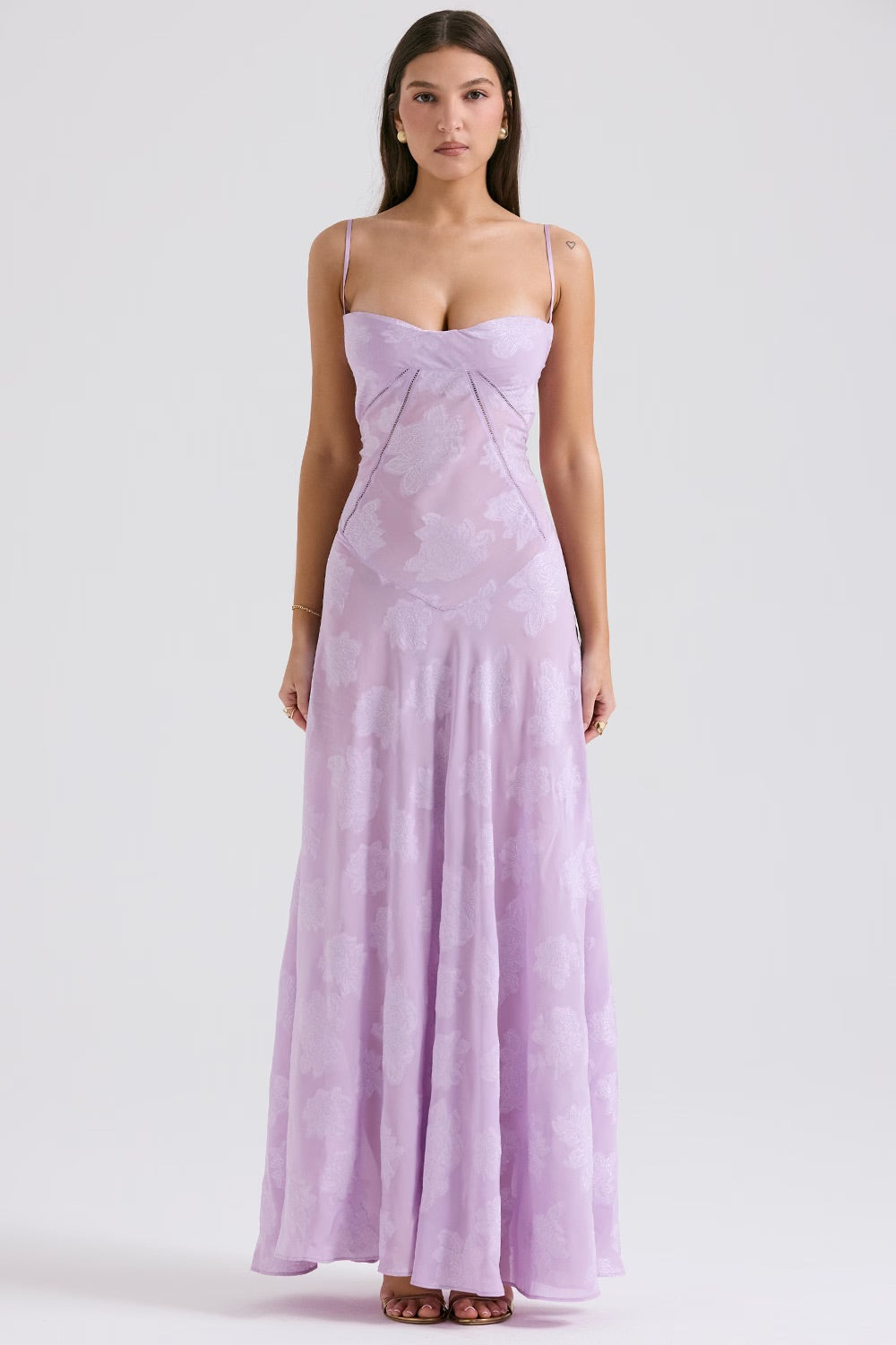 Frau in langem, fliederfarbenem Abendkleid mit Spaghettiträgern und floralem Muster, stehend vor weißem Hintergrund. Elegantes Damenmode-Outfit.
