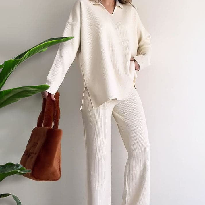 Frau in cremefarbenem Strickset mit Pullover und Hose, hält braune Plüsch-Tasche, neben grüner Pflanze. Modetrend, gemütlicher Look, Herbstmode.