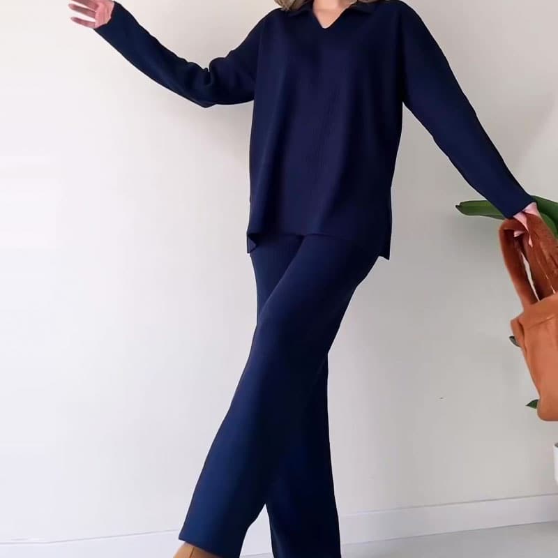Dame in dunkelblauem Strickanzug posiert vor weißer Wand, hält braune Tasche. Modetrend, eleganter Freizeitlook, Herbstmode 2023.