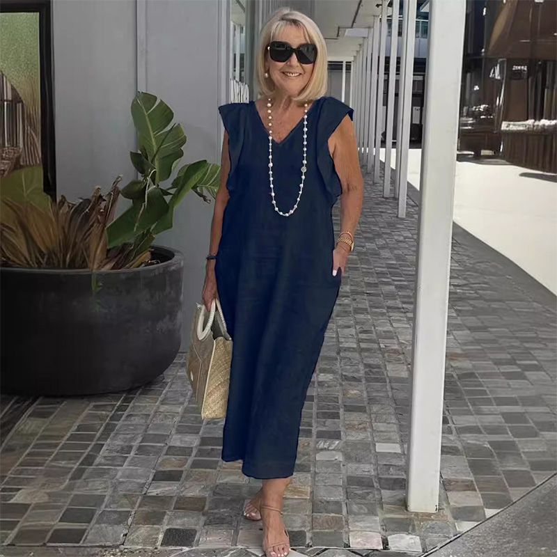 Ältere Frau in dunkelblauem Sommerkleid mit Perlenkette und Sonnenbrille, hält eine Strohtasche, steht auf gepflastertem Gehweg neben Pflanze.