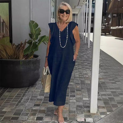 Ältere Frau in dunkelblauem Sommerkleid mit Perlenkette und Sonnenbrille, hält eine Strohtasche, steht auf gepflastertem Gehweg neben Pflanze.