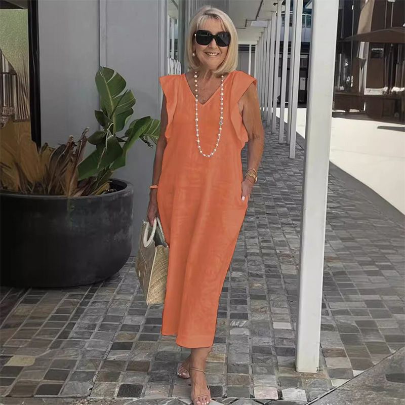 Ältere Frau in orangefarbenem Sommerkleid, Sonnenbrille und Perlenkette, spaziert auf gepflastertem Weg. Elegante Mode für Damen über 50.