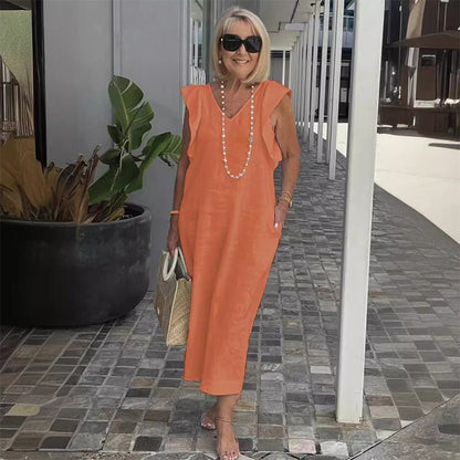 Ältere Frau in orangefarbenem Sommerkleid, Sonnenbrille und Perlenkette, spaziert auf gepflastertem Weg. Elegante Mode für Damen über 50.