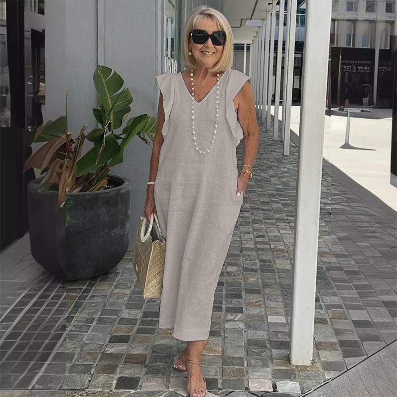 Ältere Frau in grauem Leinenkleid mit Perlenkette, Sonnenbrille und Strohtasche, steht lächelnd auf gepflastertem Gehweg neben Pflanze.