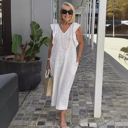 Ältere Frau in weißem Sommerkleid mit Sonnenbrille und Strohtasche, steht auf gepflastertem Weg neben großen Pflanzen. Elegante Mode für Damen.
