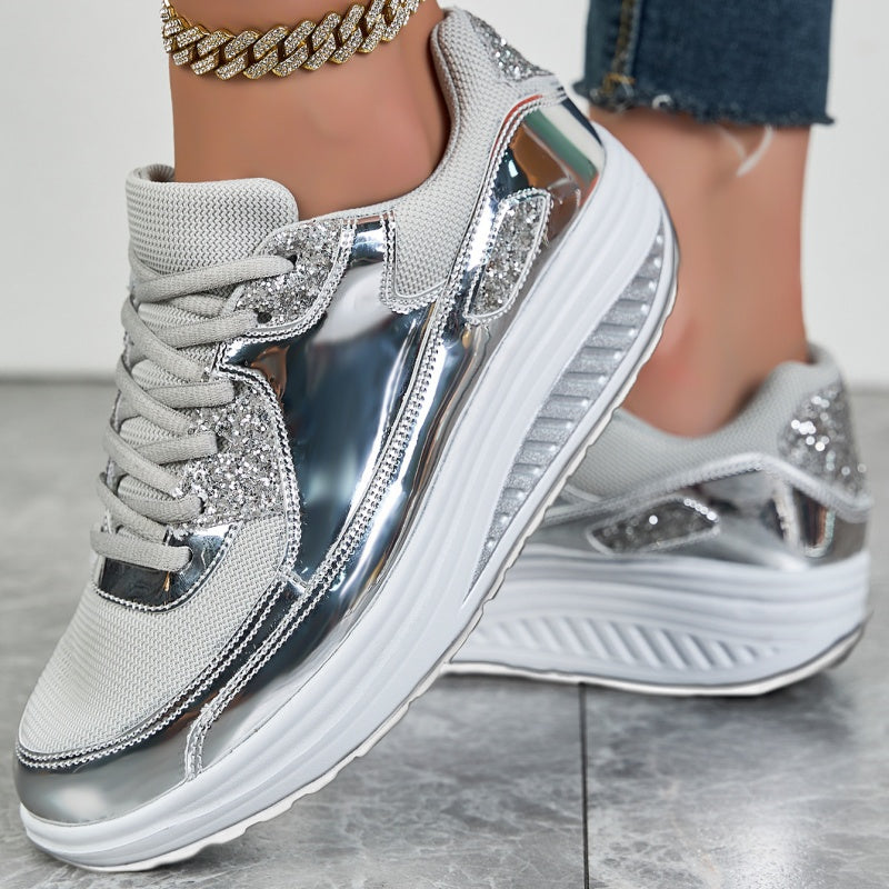 Silberne Damen-Sneaker mit Glitzerakzenten und dicker Sohle, kombiniert mit Jeans und goldener Fußkette. Modische Sportschuhe für Damen.