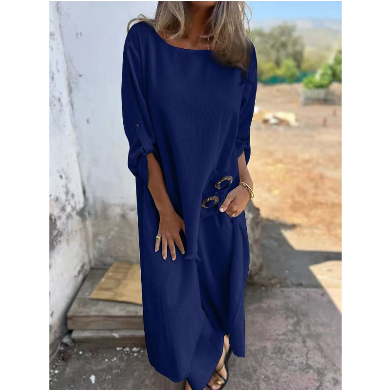 Frau in blauem Leinenkleid, hält Sonnenbrille, steht draußen vor weißer Wand. Sommermode, lässiger Stil, elegantes Freizeitoutfit.