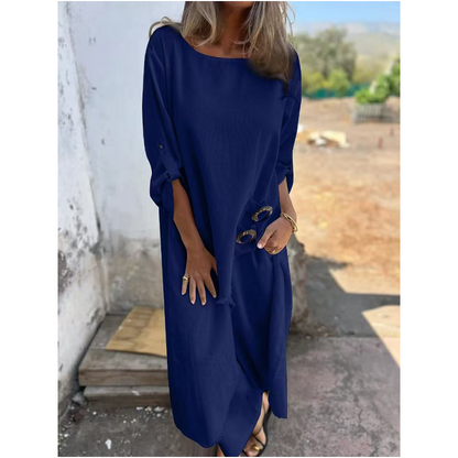 Frau in blauem Leinenkleid, hält Sonnenbrille, steht draußen vor weißer Wand. Sommermode, lässiger Stil, elegantes Freizeitoutfit.